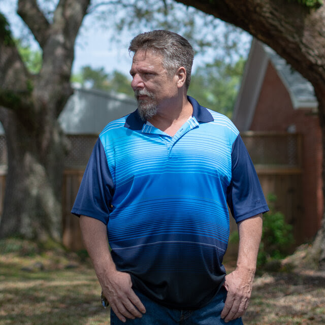 John Barnett is wearing a blue short-sleeved shirt, standing in an area with trees while he places his hands in his pockets.