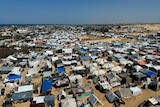 An aerial view of hundreds of tents set up on brown sand across a stretch of land with a blue sky in the distance.