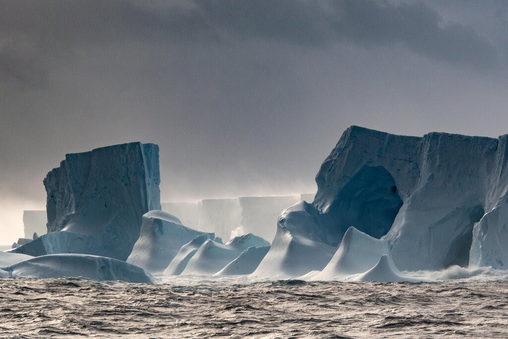 The edge of an iceberg shows signs of erosion and melting, under a gray cloudy sky.