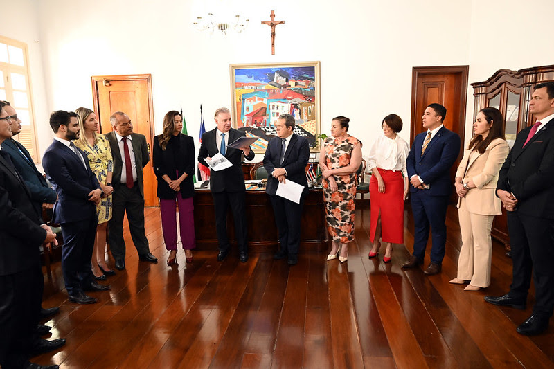 Foto horizontal de lançamento do Plano de Ação de Alcântara, por TJMA e parceiros. A imagem mostra um grupo de pessoas no Gabinete da Presidência do TJMA, todas de frente para a câmera, enquanto o presidente do Tribunal, desembargador Froz Sobrinho, fala e gesticula, enquanto segura documentos. Ao fundo, há uma grande pintura que retrata uma cena colorida de uma vila, e acima dela, há um crucifixo na parede. As paredes da sala são brancas. O piso é de madeira marrom.