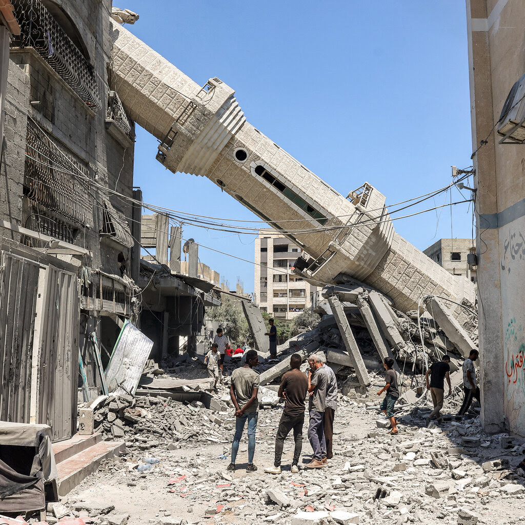 People on a street looking at a toppled minaret that is leaning on a building.