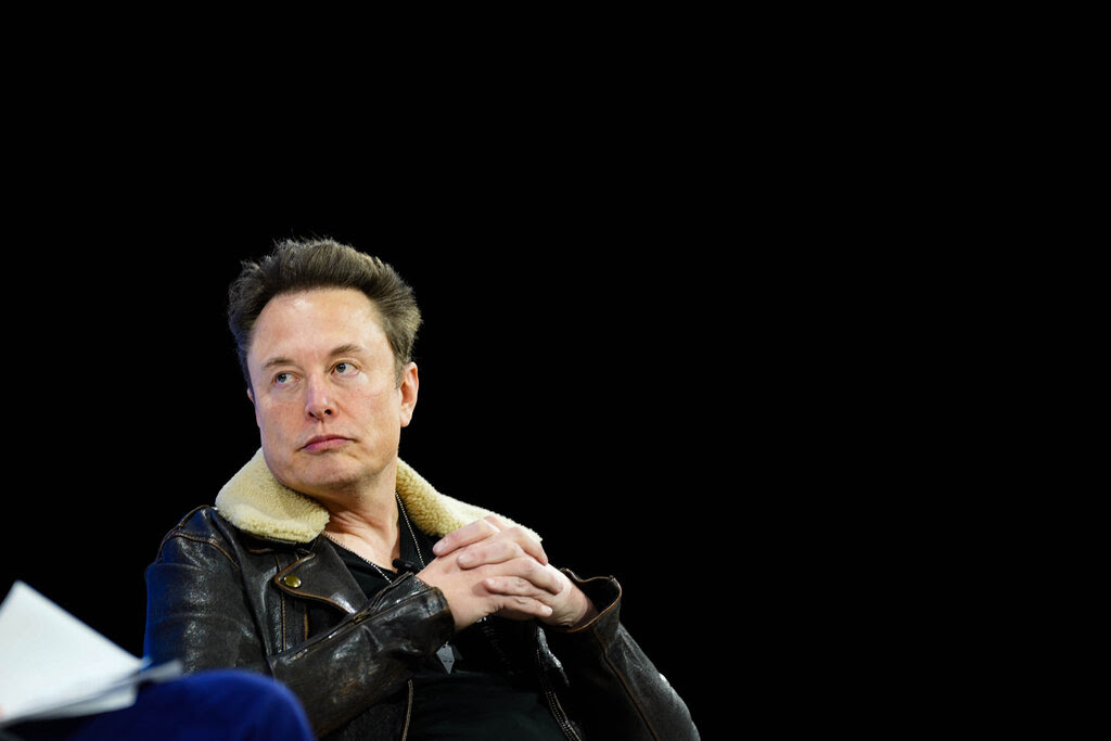 Elon Musk in a black leather jacket, his hands folded, apparently listening to someone off camera.