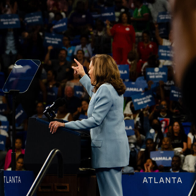 Kamala Harris speaks at a rally. A person holds up a “Kamala” sign in the foreground.