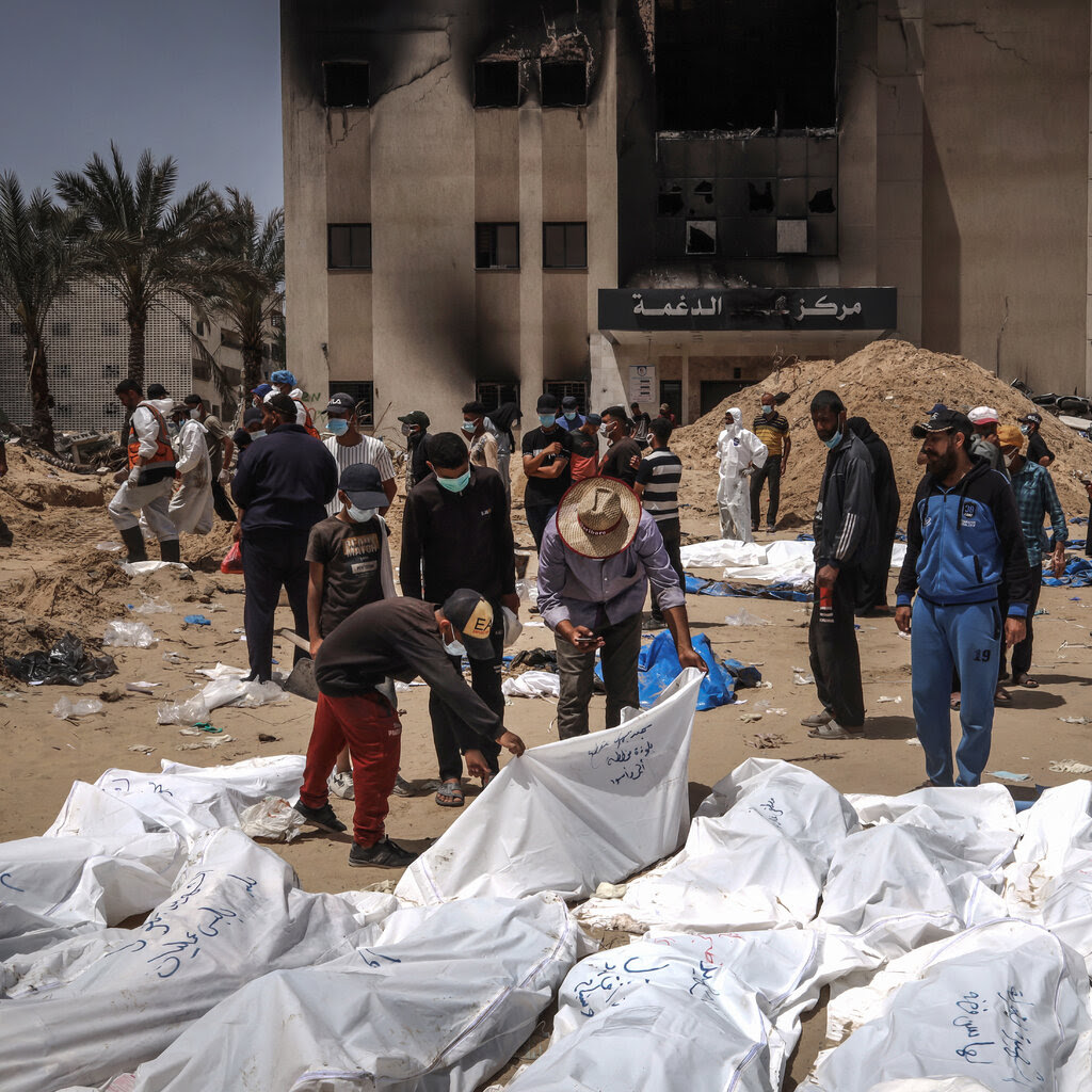 People looking at white body bags lying on sandy ground. 