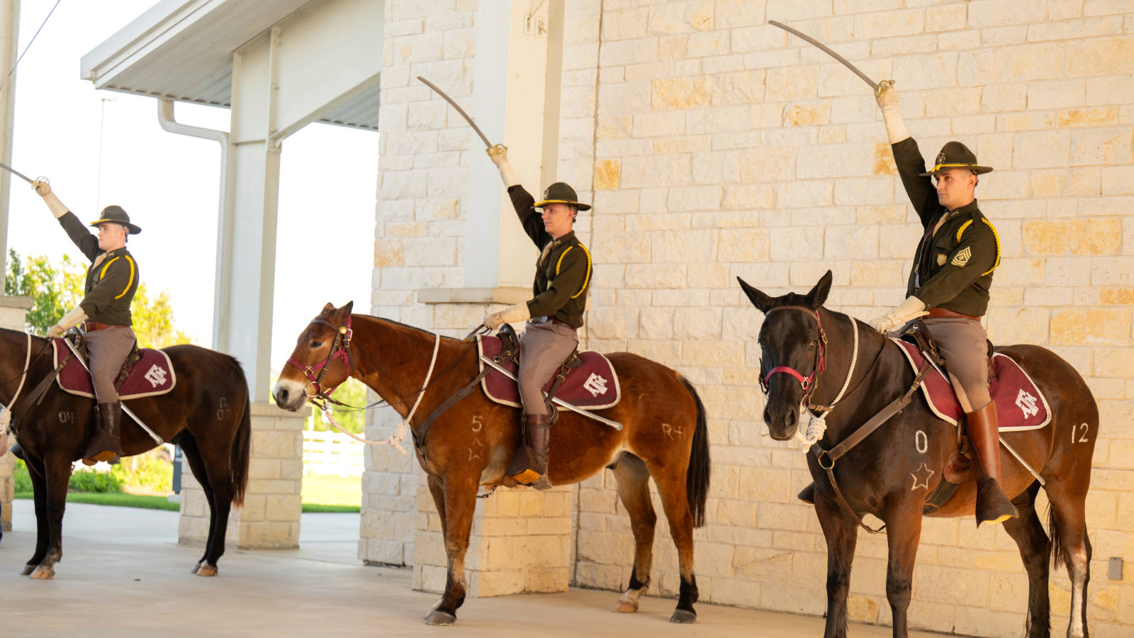 Three men with sabers raised to the sky, in uniform and wide brim military hats, sitting on horses with Texas A&M saddle blankets.