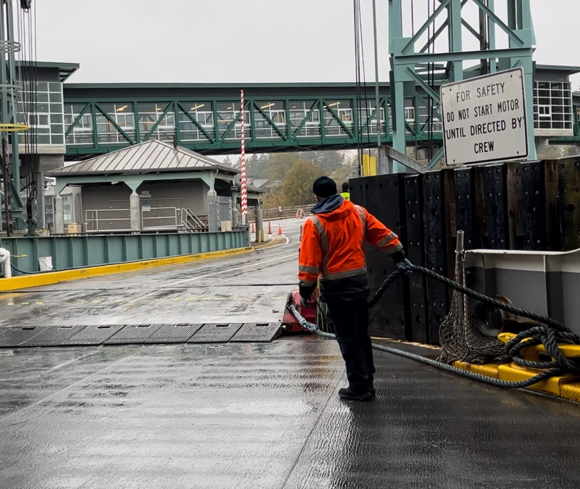 Person in an orange jacket holding a rope at a dock, with a steel walkway and safety sign in the background