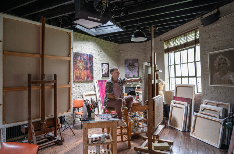 The artist Jonathan Yeo sits painting before a canvas on an easel in his studio. Stacks of canvases lean against the wall on the floor. 