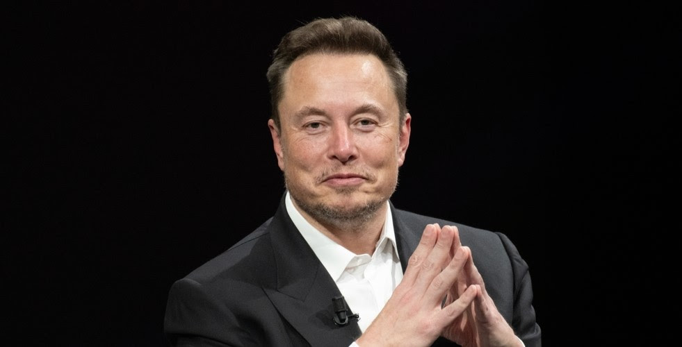 Elon Musk, vêtu d'une veste noire et d'une chemise blanche, sourit à la caméra et tient ses mains de manière à ce que les extrémités de ses doigts se touchent.