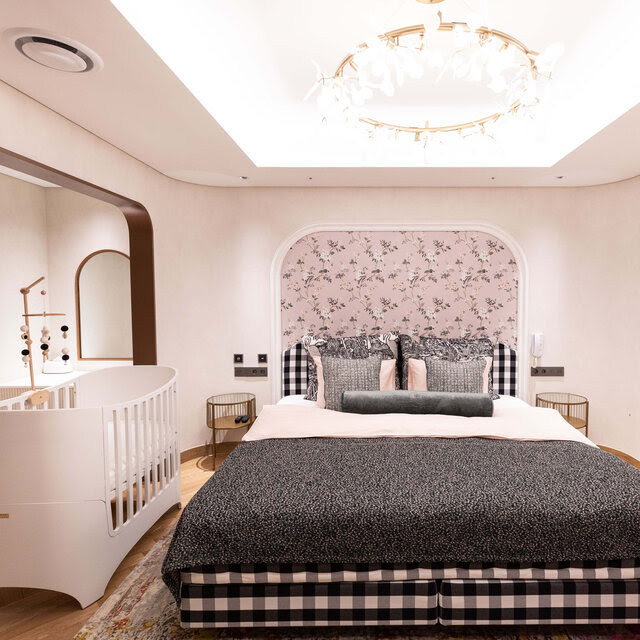 A windowless but luxurious hotel-like room with a large bed and a baby’s crib next to it.