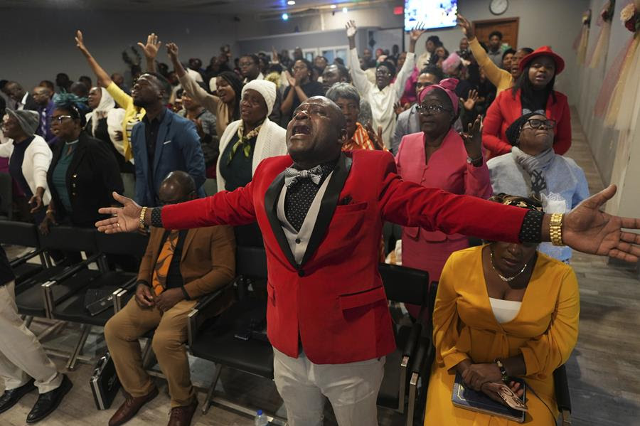 A Black man dressed in formal clothing stands with his arms outstretched and head tipped back in prayer. Behind him are other people gathered in the church.