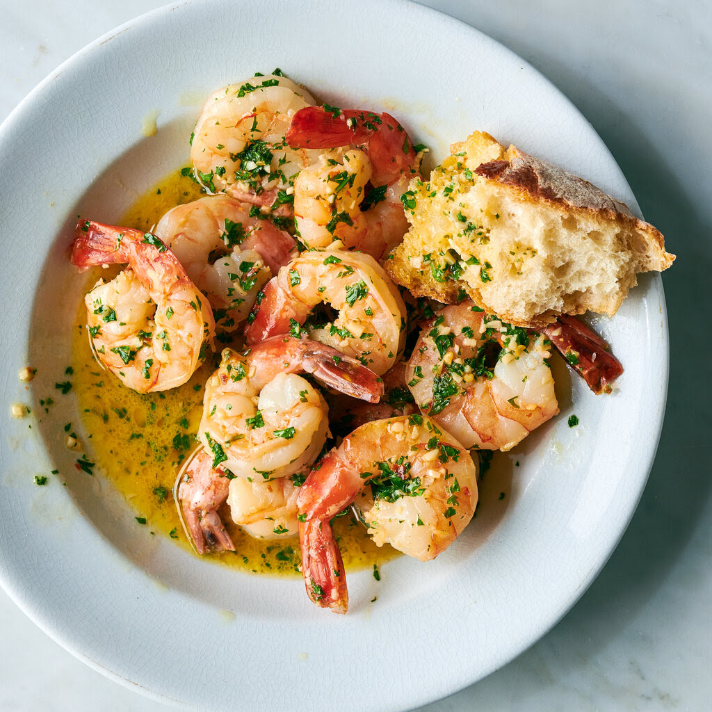 A simple white plate holds a serving of buttery shrimp scampi with a torn hunk of bread for soaking up the briny, winy juices.