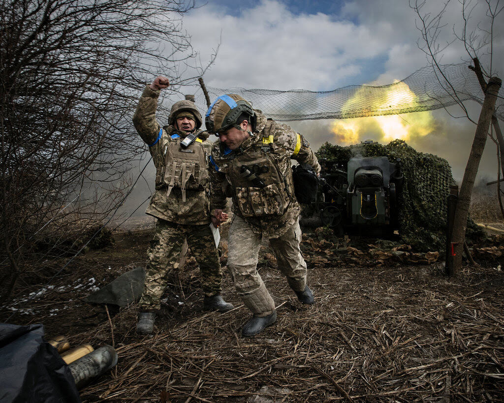 Two soldiers moving away from a howitzer that is firing artillery rounds behind them.