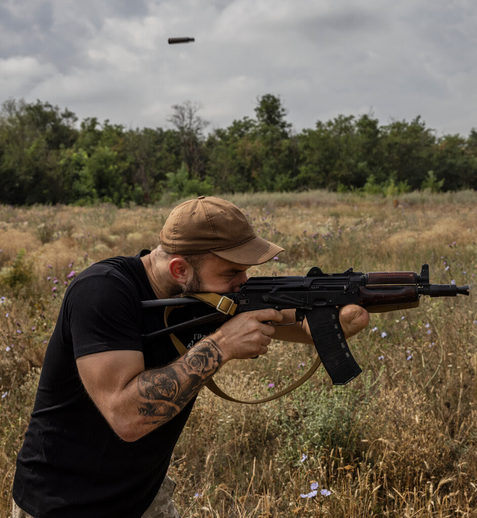 A man in a black T-shirt pointing an assault rifle in a field. It rests on the stump that now terminates his left arm.