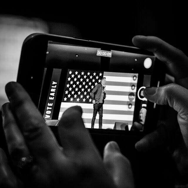 A phone records a video of Elon Musk speaking in front of an American flag.