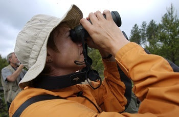 a woman with short, dark hair and wearing a tan, floppy hat and orange jacket looks through black binoculars