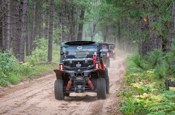 three black and red off-road vehicles drive single-file down a dirt trail in a heavily forested area
