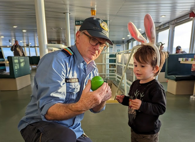 Ferry crewmember holding a plastic Easter egg as a child wearing bunny ears looks on while inside the passenger cabin