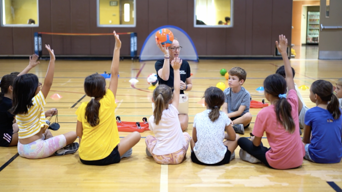 Group of children sitting on a gym floor in a semi-circle raising their hands as teacher sits in front of them holding up a volleyball