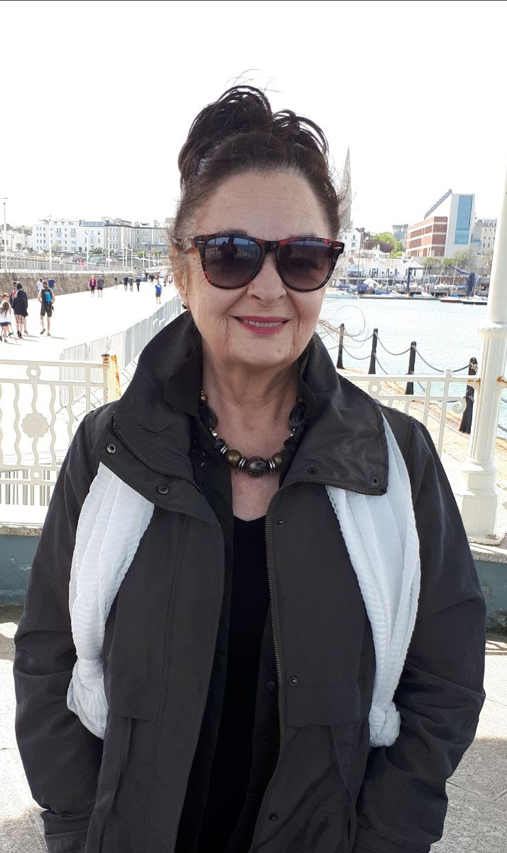 Writer Celia de Fréine stands on a harbour while wearing a coat and sunglasses.