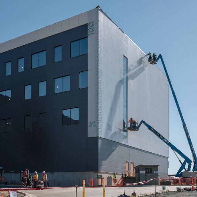 Workers on blue cranes at a construction site work on a building with several windows, with construction equipment on a dirt area in the foreground.