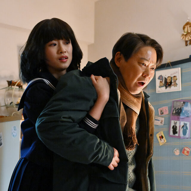 A young Japanese woman stands behind her father, appearing to hold him up or back. There are many small decorations on the walls in the background. 