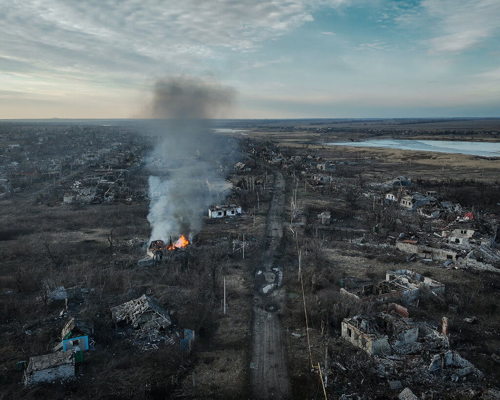 An aerial view of a ruined village. Smoke is rising from a house that is on fire.