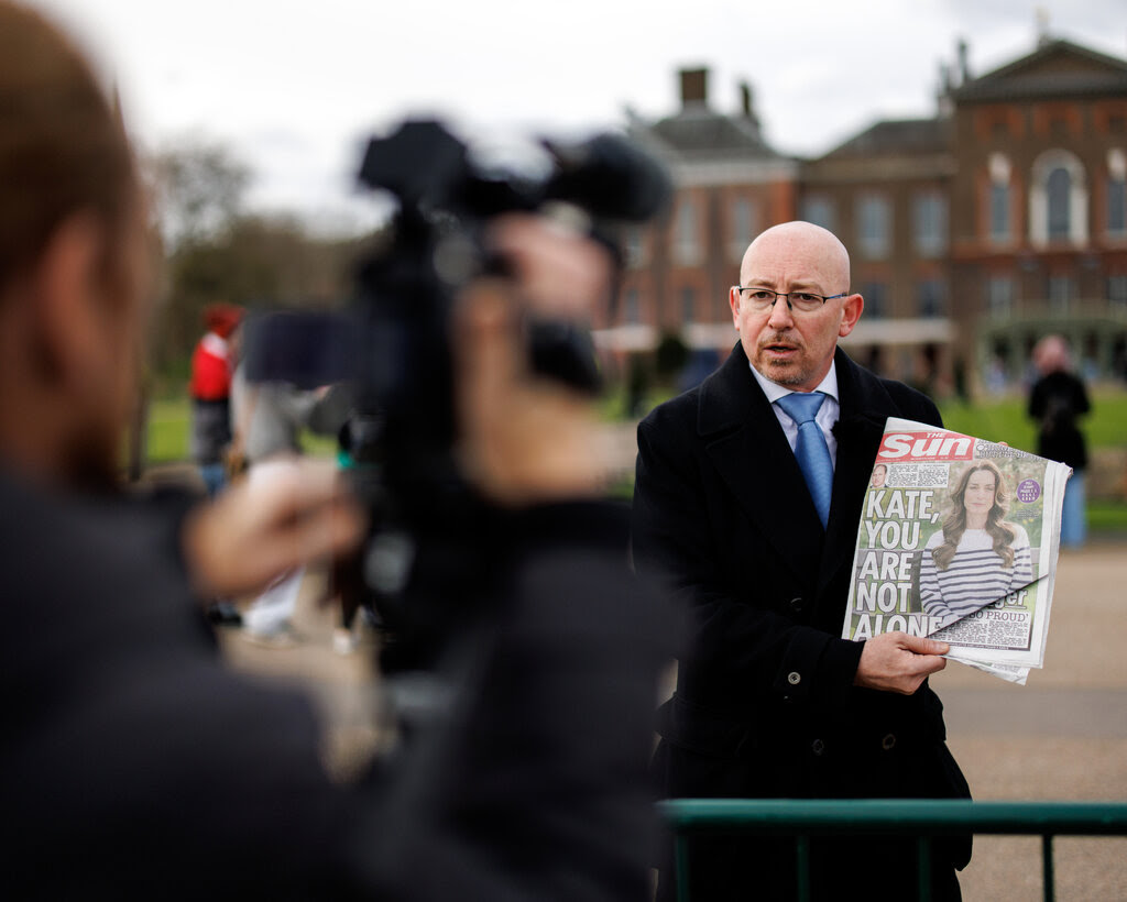 A man in a dark overcoat and blue tie holds up a copy of The Sun newspaper outside Kensington Palace in London.