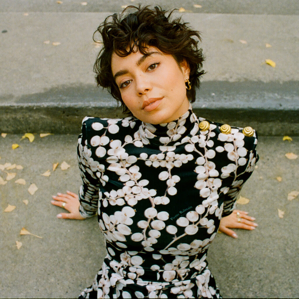 Auli’i Cravalho, in a patterned black-and-white dress, leans back with her palms propping her up on a concrete step.