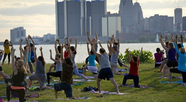 About two dozen women and men in exercise gear do yoga poses on shoreline along Detroit river. Buildings frame the skyline.
