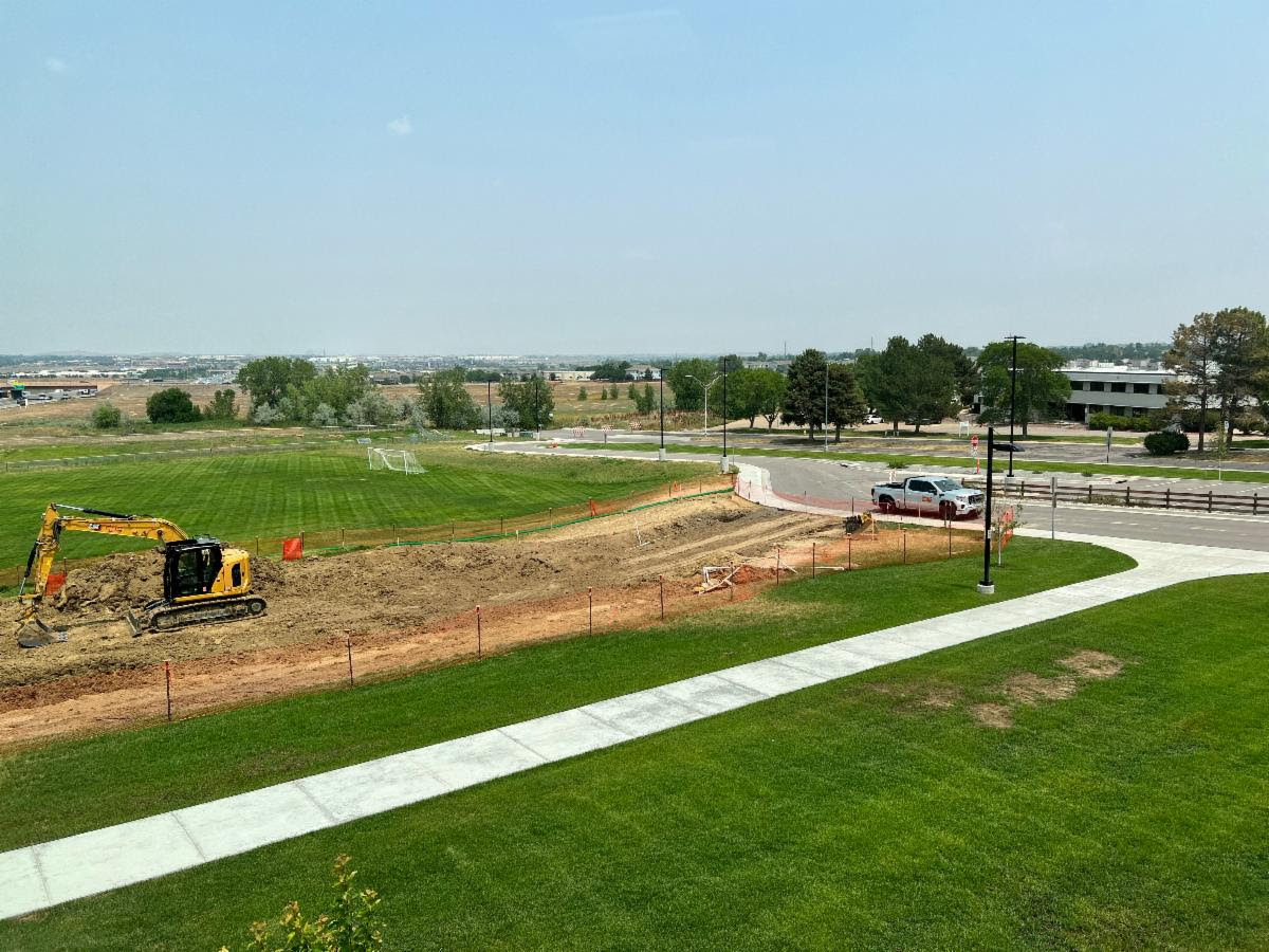 Construction site with a yellow excavator near a green lawn and a white truck on a road