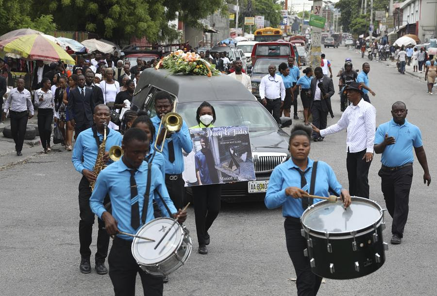 A funeral procession for Judes Montis, a mission director who was killed by gang members. There are people with various instruments walking in front of the hearse.