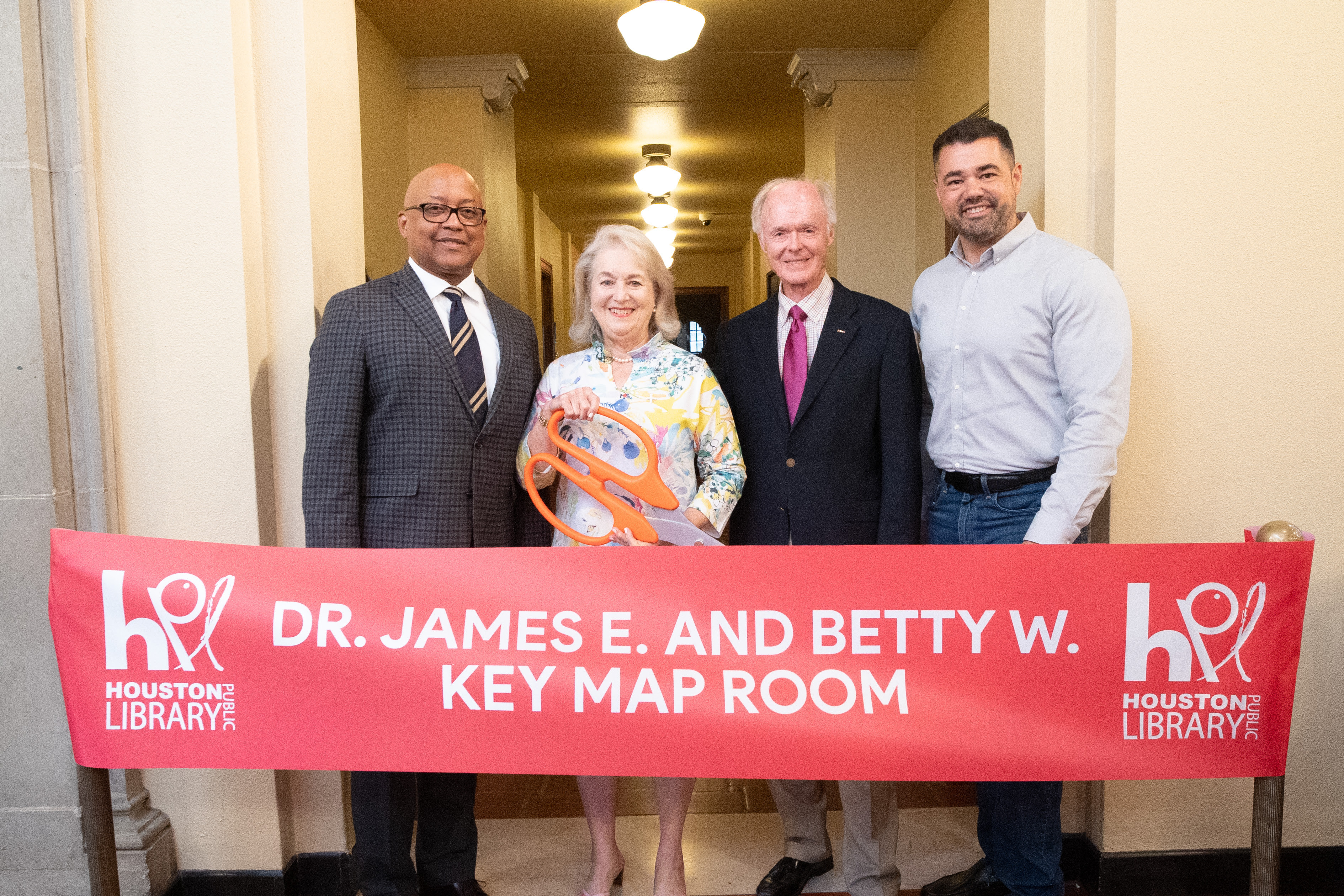Three men, one woman smiling and standing side by side at a ribbon cutting ceremony. The woman in the center is holding large, orange ceremonial scissors, poised to cut a wide ribbon.