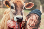 Nellie Geraghty smiling while standing next to a brown cow. Nellie wears a beanie and red patterned sweater with overalls.