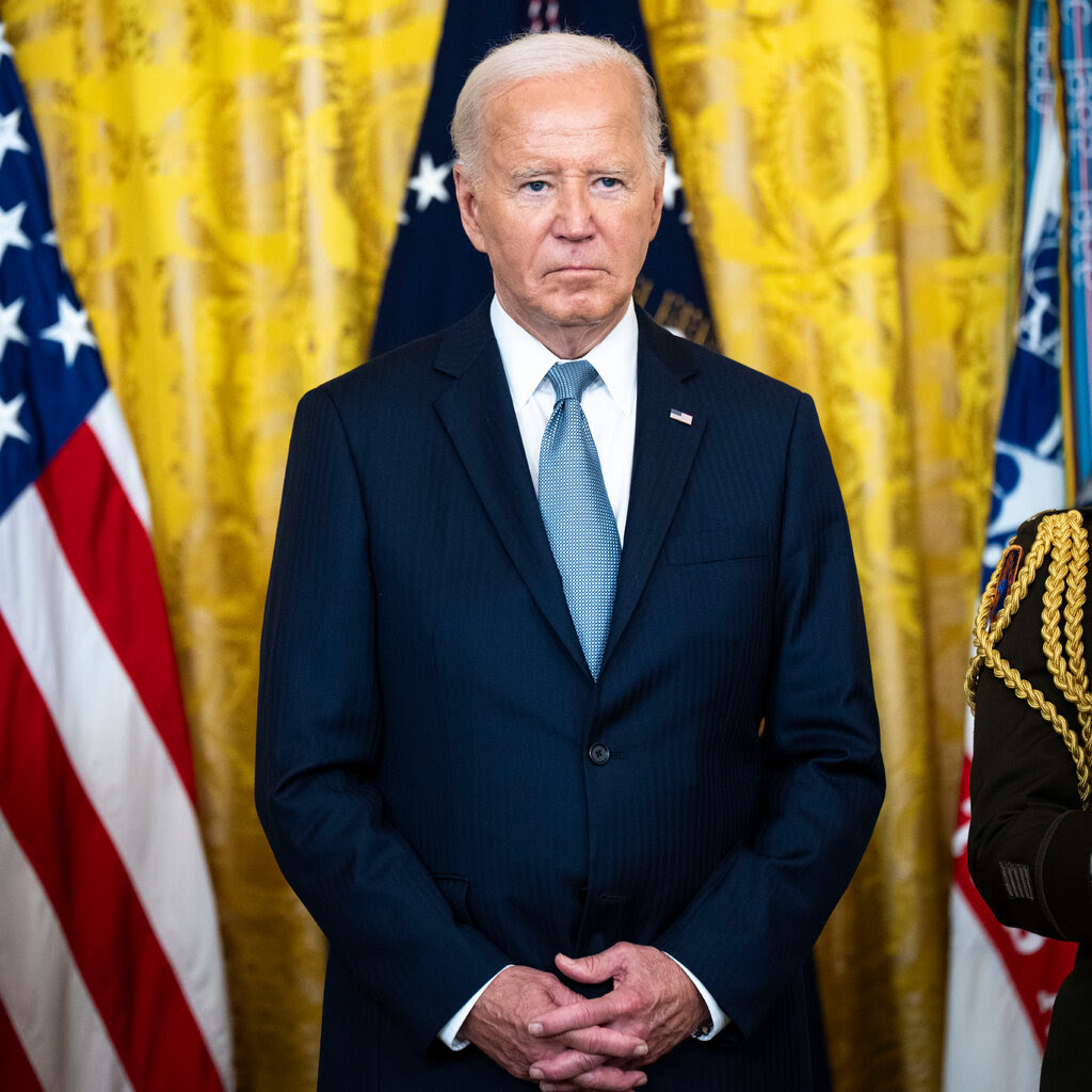 President Biden in a blue suit and tie, standing in front of several flags.