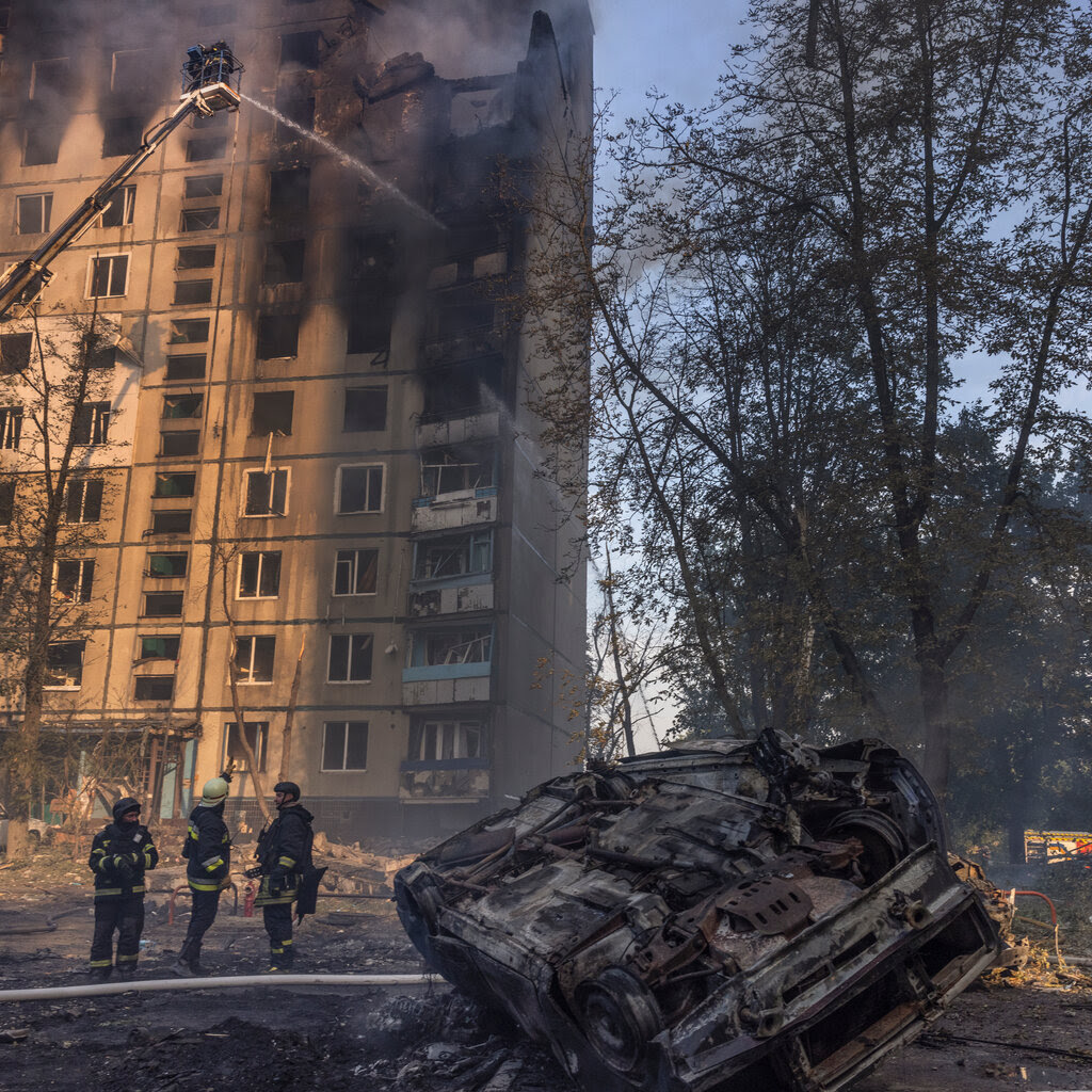 Firefighters stand in front of a burning multistory building. A burned and overturned vehicle is beside them. 