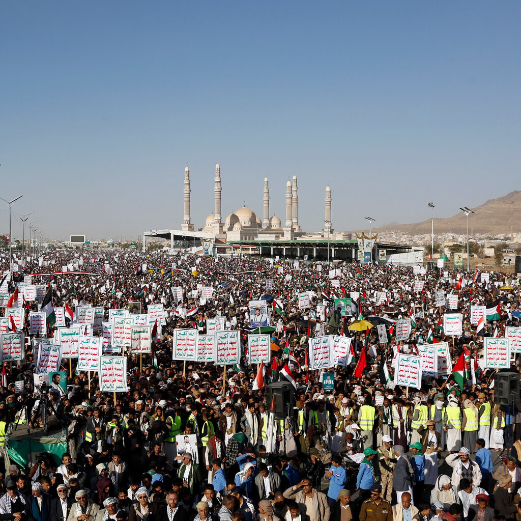 A large crowd, many holding up signs in Arabic, march on a street. There is a mosque in the background and a hill.