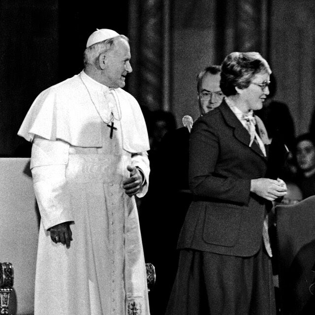 A black-and-white photo of Pope John Paul II standing on a stage next to a nun in a skirt and blazer.