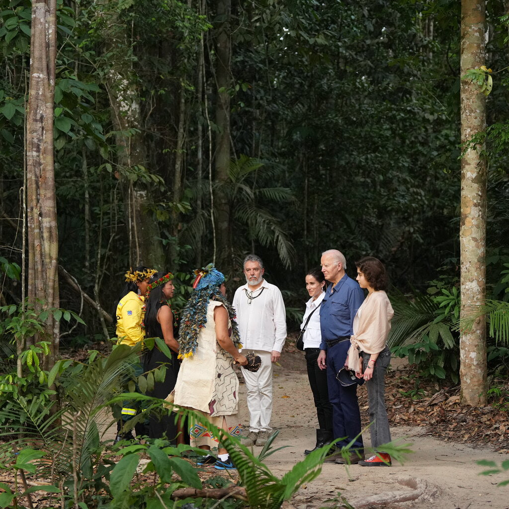 President Biden, in a blue shirt, meets with three Indigenous people in a forest.