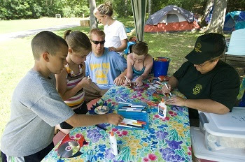 A woman volunteering as a campground host helps young campers at a picnic table work on a nature-themed arts and crafts project.