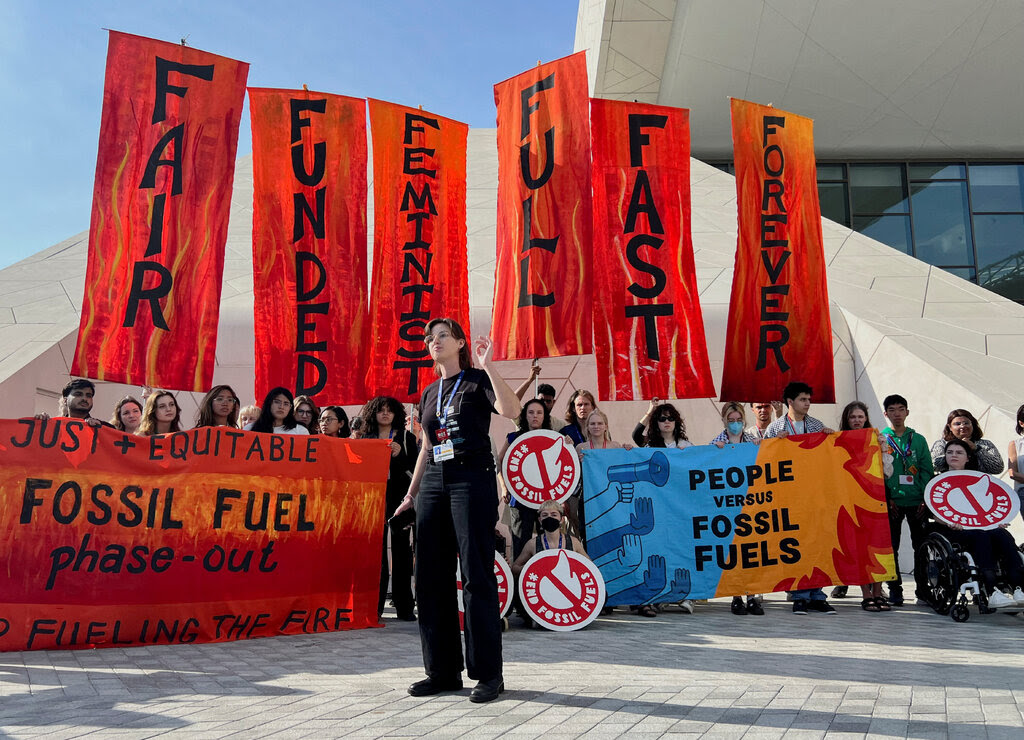 About two dozen protesters standing outside a conference center with banners. Six of the banners, lined up side-by-side on tall poles and referring to the global energy transition, read “fair, funded, feminist, full, fast, forever.”