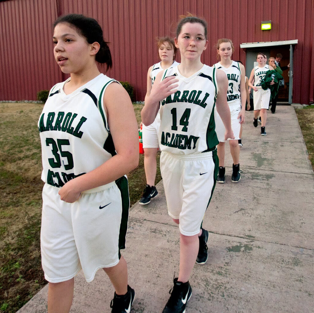 A line of teenage girls in white basketball uniforms walks out of a red building.