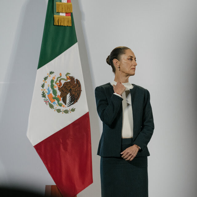 Claudia Sheinbaum, con traje oscuro, delante de una bandera mexicana.