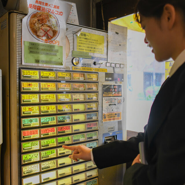 A woman with her hair in a ponytail points to a bright green button on a machine filled with rows of colorful buttons with Japanese writing.