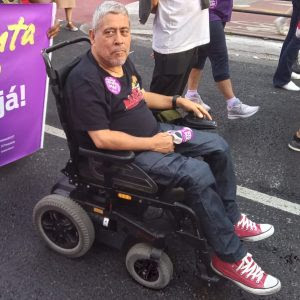 A imagem mostra a foto de Isaias Dias, que est sentado em uma cadeira de rodas motorizada, usando uma camiseta preta e tnis vermelhos. Ele est em um ambiente externo, possivelmente em um evento ou manifestao, com pessoas ao fundo