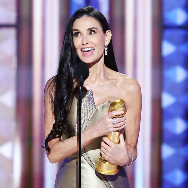 A woman with long brunette hair stands at a microphone smiling and holding a Golden Globe Award.