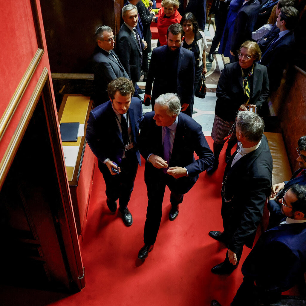 Several people in suits walk down a hallway carpeted in red.