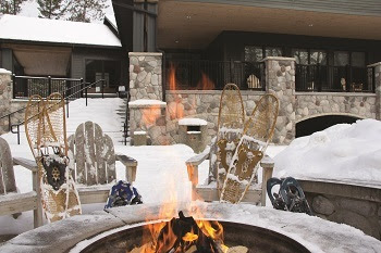 snow-dusted chairs and snowshoes around a gray concrete firepit in front of a fieldstone and steel lodge-style building