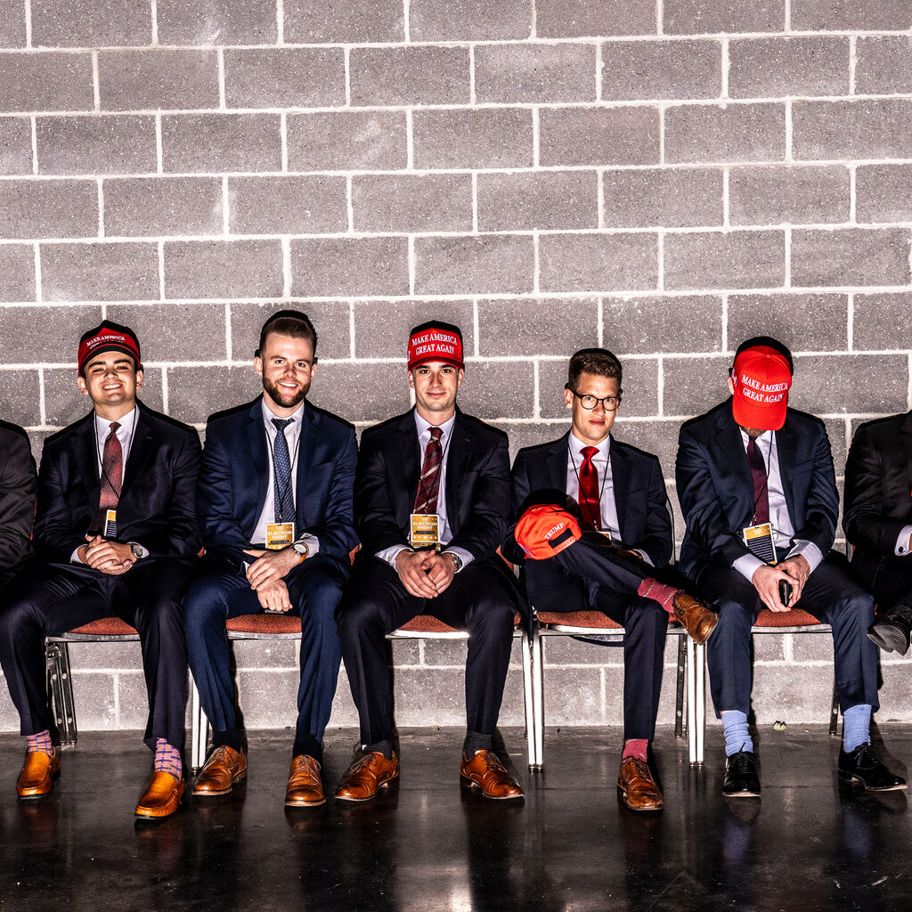 A group of suited young men, some wearing “Make America Great Again” baseball hats, are seated in chairs against a brick wall.