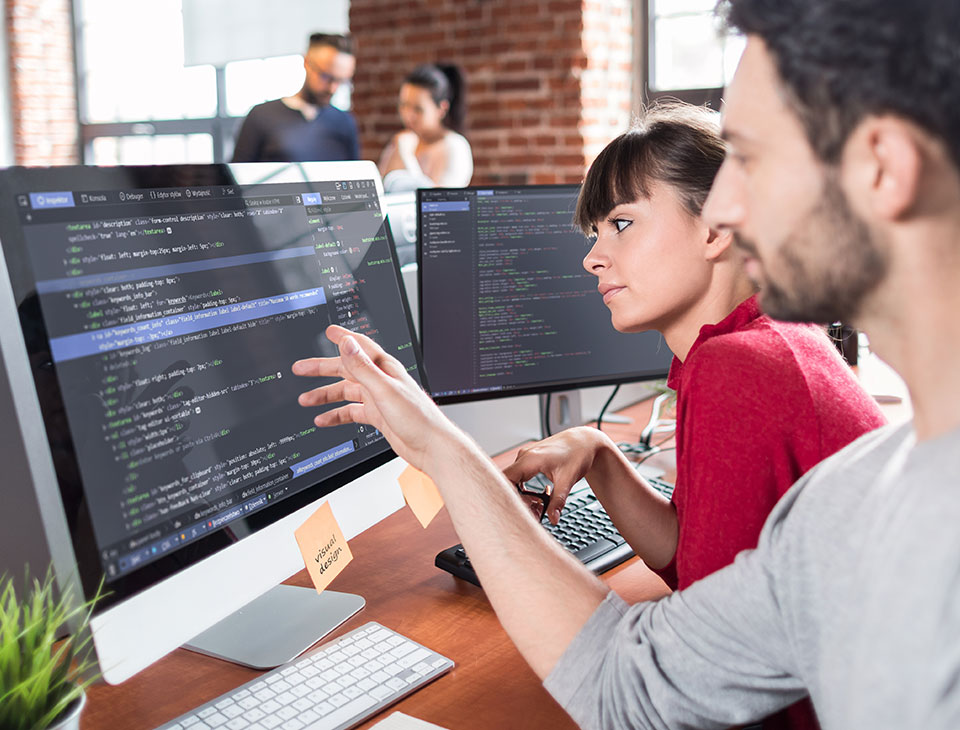 A man and a woman are seated together, looking at a computer screen displaying data. They appear focused and engaged, possibly analyzing information or collaborating on a project. The computer screen illuminates their faces as they study the data, conveying a sense of concentration and teamwork in a professional setting.