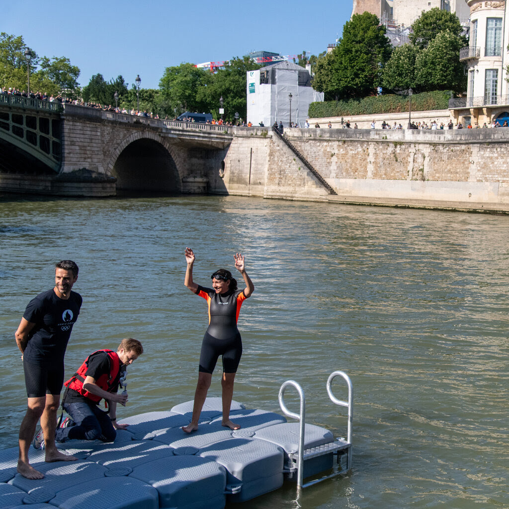 A woman standing on a floating platform in a river raises her arms.